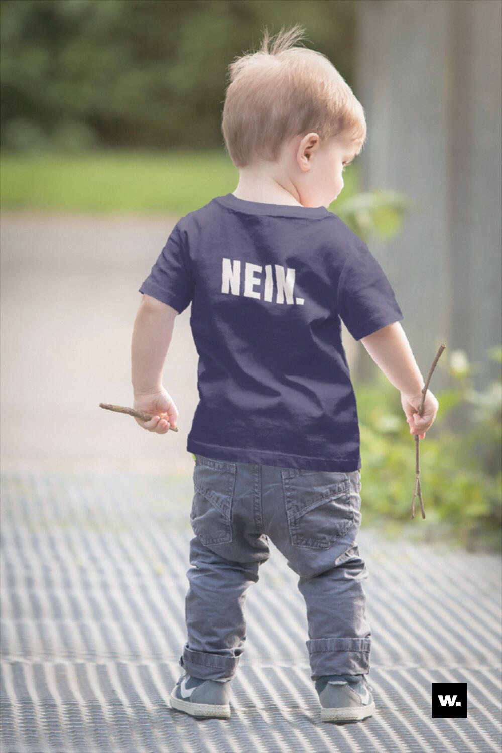 Kleines Kind von hinten in einem dunklen T-Shirt. Auf dem Rücken steht groß und weiß „NEIN.“. Das Kind hält in beiden Händen kleine Stöcke und steht draußen auf einem Weg.